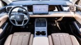 Interior of a luxury car with a wide central touchscreen on the dash, beige perforated seats, and a dark steering wheel with silver accents.