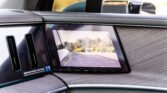 Close-up of a car interior showing a large center touchscreen displaying a rear parking camera view over an empty lot with palm trees in the distance.