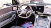 Modern car interior with a central steering wheel, large touchscreen displays, and wood trim along the dashboard during daylight outside the windows.