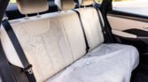Beige leather rear car seats with three headrests, black seat belts, and a protective gray sheet covering the seat area inside a car.