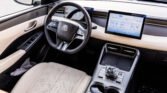 Luxury car interior with a leather steering wheel, beige perforated seats, and a wide tablet-style dashboard display on the passenger side center console.