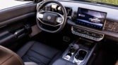 Inside a luxury car: steering wheel with logo, digital instrument cluster, and a wide touchscreen displaying an SUV image.