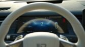 Interior view of a car cockpit featuring a large digital dashboard showing 0 km/h and vehicle status icons, with a steering wheel in the foreground