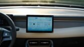 Car interior with a central touchscreen showing a blue app grid, steering wheel on the left and dashboard vents visible.