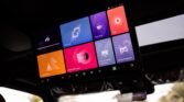 In-car infotainment touchscreen mounted on the roof console, displaying colorful app tiles like Nap Mode and Multimeda Center for vehicle controls (informational image).