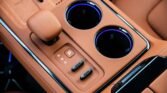 Tan leather car center console with gear selector in Park and two blue-lit cup holders nearby.