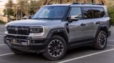 Gray SUV parked in a lot, showing front-left view with rugged bumper and large wheels against a backdrop of palm trees and buildings.