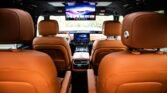Rear-seat perspective of a luxury SUV with tan leather seats, a center console, and ceiling and dashboard infotainment screens displaying content.