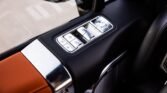 Car door control panel with silver window and mirror buttons on a black armrest, adjacent to tan leather interior