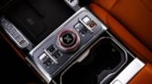 Center console of a luxury car with a large circular rotary dial surrounded by chrome, flanked by small buttons and orange leather seats in the background.