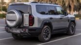 Rear view of a large gray SUV with a spare tire mounted on the back, parked in a lot with palm trees in the background.