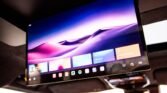 In-car ceiling monitor showing a colorful purple desert wallpaper and app tiles for entertainment in a vehicle interior