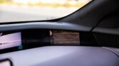 Interior of a car: dashboard and screens with a small rear‑view mirror showing a parking lot with white-lined spaces beyond the car.