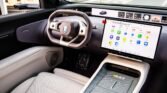 Interior of a modern car with a large horizontal touchscreen displaying app icons beside a beige steering wheel and center console.