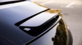 Close-up of a glossy black car trunk with a rear spoiler reflecting the surroundings in a parking lot.