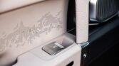 Close-up of a luxury car interior: beige panel with a chrome release button labeled OPEN and a textured white armrest beside a speaker grille.