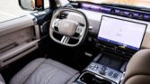 Interior of a luxury SUV cabin with beige seats, a wrapped steering wheel, and a large horizontal touchscreen dashboard visible across the front.