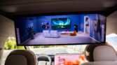 Large in-car display showing a blue living-room setup with white sofas, a wall TV, and plants in the scene