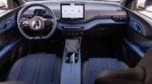 Interior of a modern car showing a blue and black dashboard, steering wheel with controls, a large central touchscreen, and two front seats with blue upholstery; center console with cup holders visible between seats.