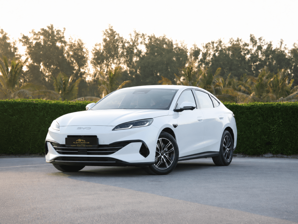 White four-door sedan parked on a paved surface with a green hedge and trees in the background.